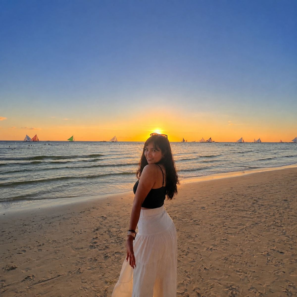 Demae Pearl Francisco, founder of MultiTasker, standing on a beach at sunset, turning to face the camera, with sailing boats on the water behind her.